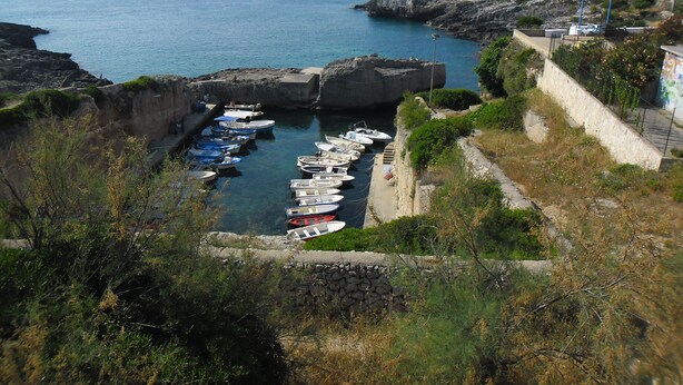 A coastal marina with boats docked, rocky cliffs, and a stone wall.