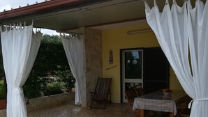 A covered patio area with stone flooring, white curtains, a wooden table with a patterned tablecloth, and a chair.