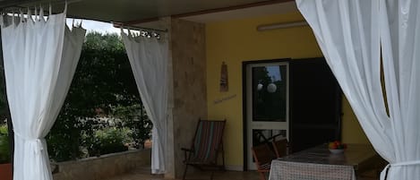 Un espacio de patio cubierto con suelo de piedra, cortinas blancas, una mesa de madera con mantel estampado y una silla.