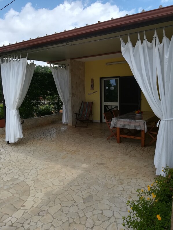 Un espacio de patio cubierto con suelo de piedra, cortinas blancas, una mesa de madera con mantel estampado y una silla.
