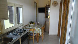 A compact kitchen with a gas stove, oven, and sink. A dining table with chairs is set up in the center, and a refrigerator is visible in the background.