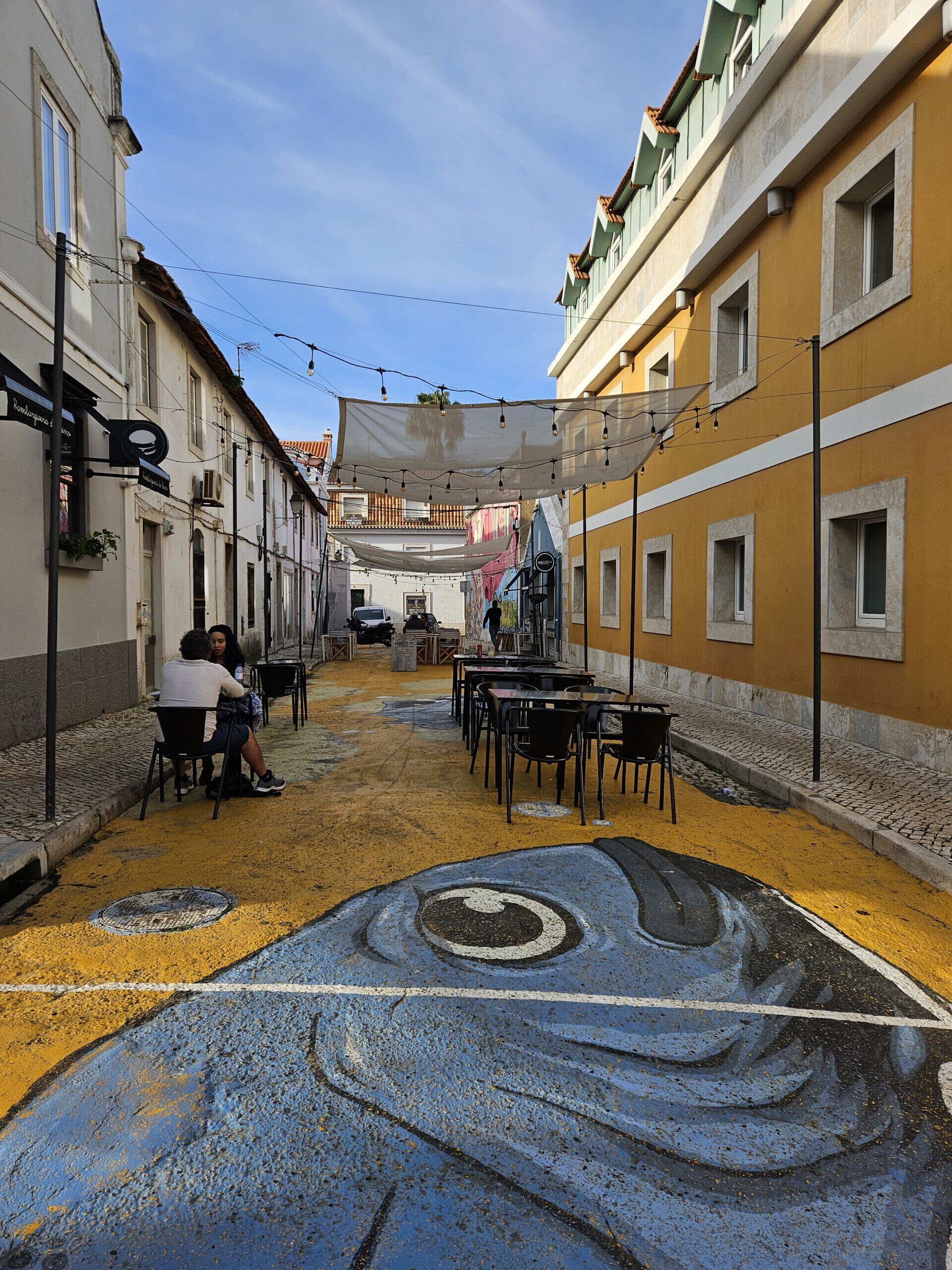 Eine schmale Straße mit Außenbestuhlung und einem Wandbild am Boden.