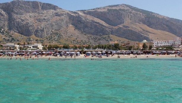 A beach with clear turquoise water, a crowd of people, and a mountainous backdrop.