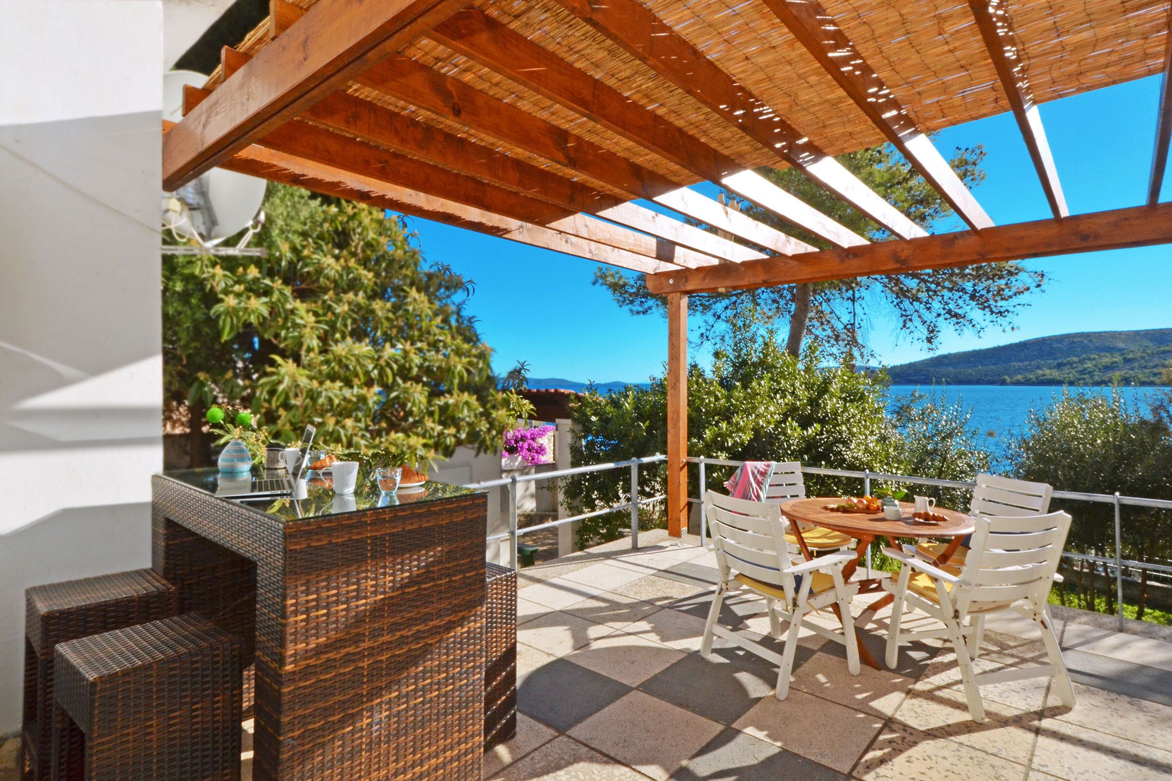 Een terras met een houten pergola, rieten meubels en uitzicht op een meer en bergen.