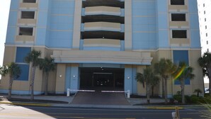 A multi-story hotel with a central entrance, balconies on each floor, and a parking area in front.