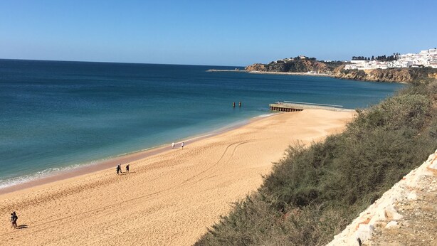 Ein Strand mit einem Steg, der sich ins Meer erstreckt, umgeben von Gebäuden auf einer Hügelside.