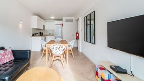 Ein modernes Wohnzimmer mit einer Couch, einem Esstisch mit Stühlen, einem flachbildfernseher an der Wand und einer kleinen Küchenzeile im Hintergrund.