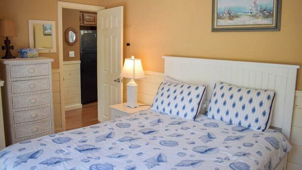 A bedroom with a bed, dresser, nightstand, lamp, and a painting on the wall.