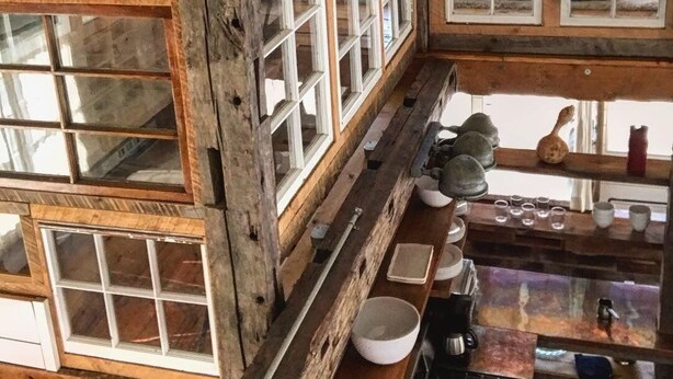 Ein rustikales Interieur mit Holzbalken, zahlreichen Fenstern und einer Küchenzeile mit Geschirr und einer Kaffeemaschine.