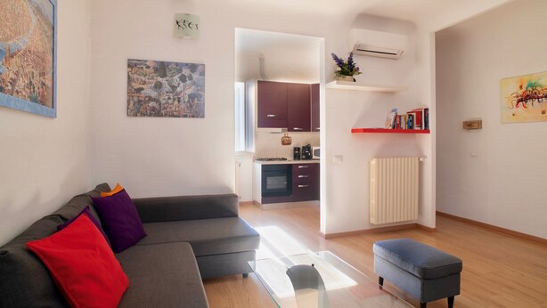 Ein modernes Wohnzimmer mit einer grauen Couch, roten und violetten Kissen, einem Glastisch und einem kleinen Hocker. Im Hintergrund ist eine Küche mit dunklen Schränken und einer weißen Arbeitsplatte zu sehen.