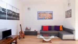 Ein Wohnzimmer mit grauem Sofa, einem Holztisch, einem Fernseher auf einem Holzschrank, einer Gitarre und einem Bild an der Wand.