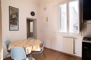 Un coin repas avec une table recouverte d’une nappe à motifs floraux, entourée de chaises bleues. Un tableau orne le mur, ainsi qu’une horloge, et une fenêtre offre une vue sur les bâtiments à l’extérieur.