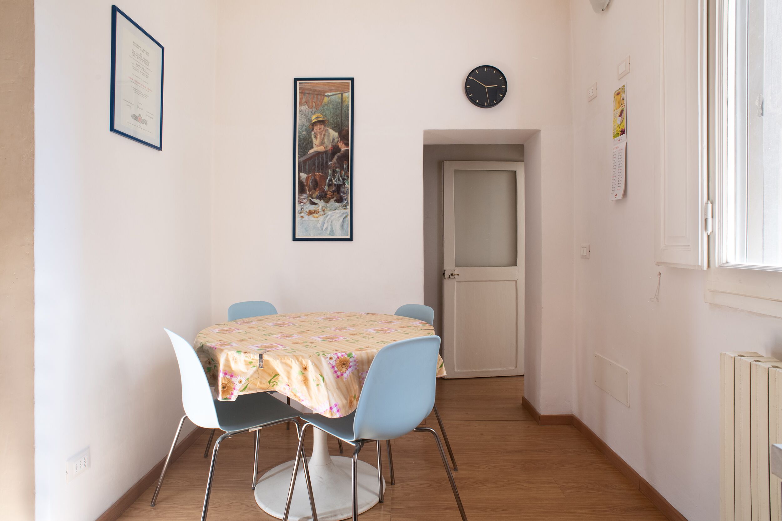 A dining area with a table covered by a floral tablecloth, surrounded by four chairs. A framed picture hangs on the wall above the table, and a clock is visible on the wall to the right.