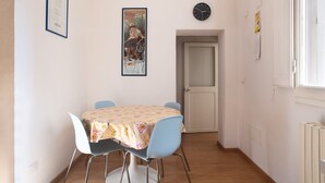 Ein Essbereich mit einem Tisch, der mit einer blumigen Tischdecke bedeckt ist, umgeben von vier Stühlen. Ein gerahmtes Bild hängt an der Wand über dem Tisch, und eine Uhr ist an der rechten Wand zu sehen.