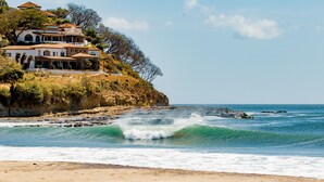 A coastal property with a large house overlooking the sea, featuring a rocky shoreline and waves crashing onto the beach. - Costa Oeste (Popoyo)