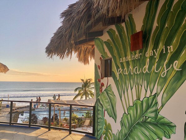 A beachside view with a pool, palm trees, and a mural with the text "another day in paradise". - Costa Oeste (Popoyo)