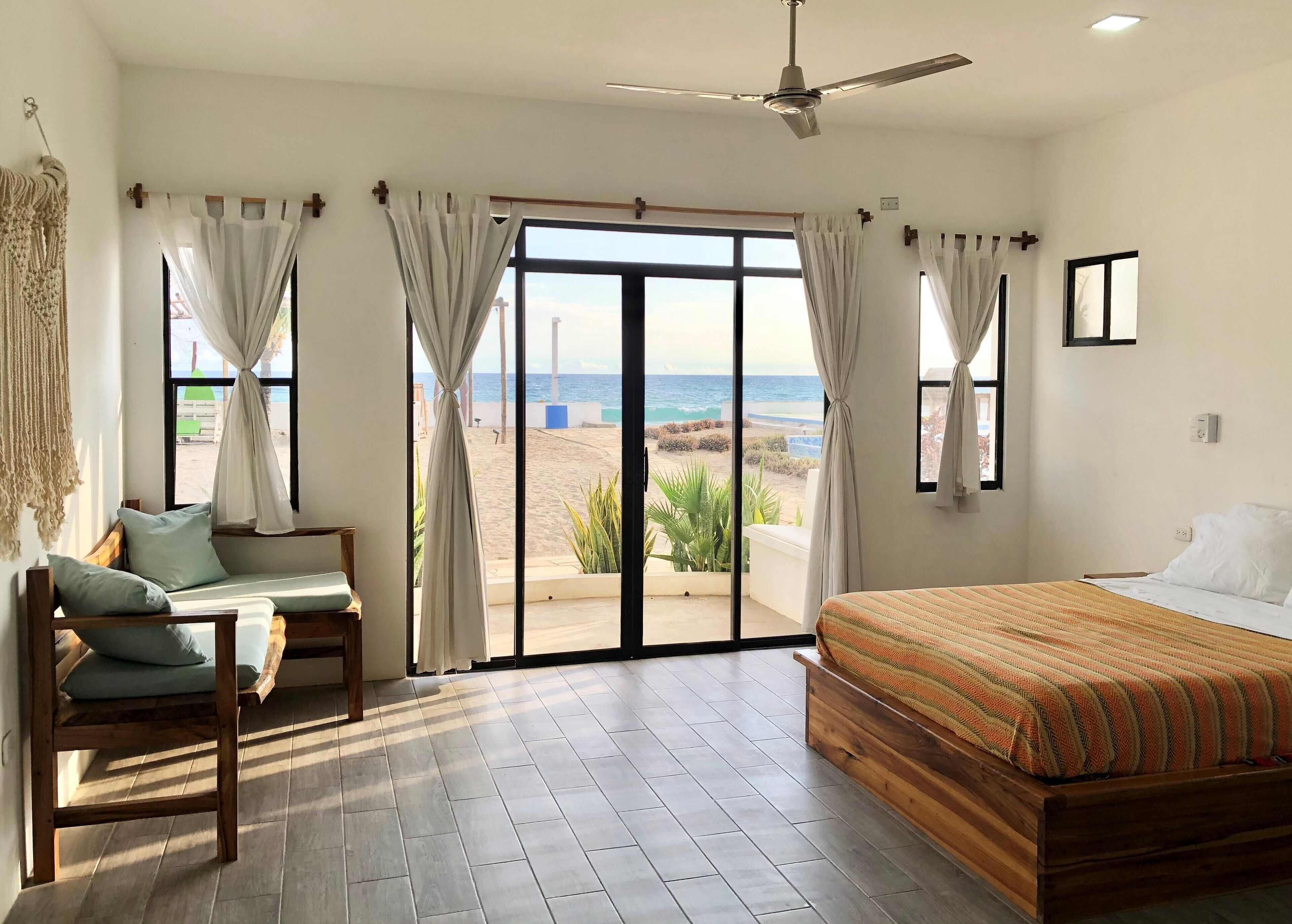 A bedroom with a bed, a seating area with a bench and cushions, and a view of the beach through large sliding doors.