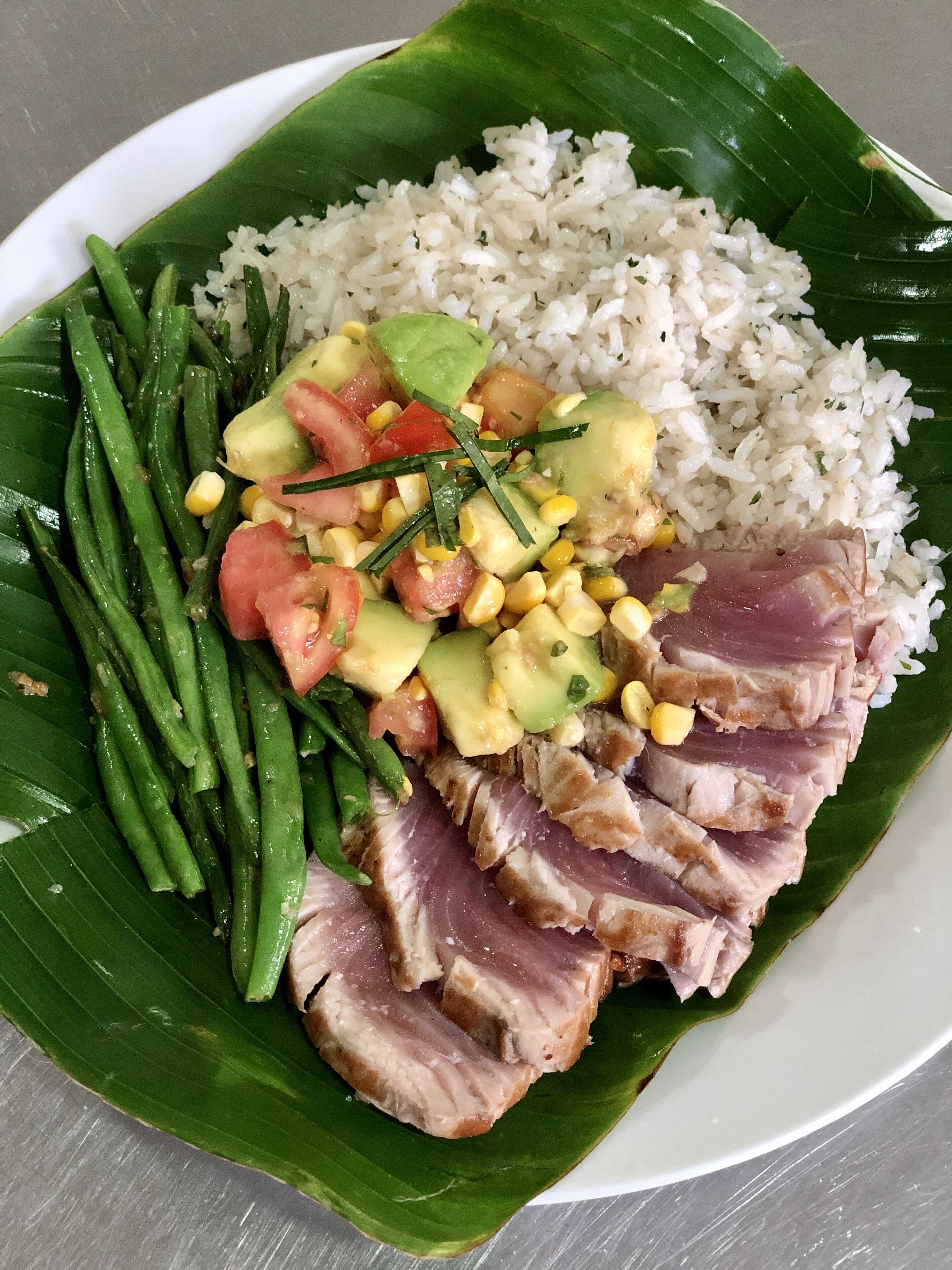 Prato de comida com arroz, vagem, abacate, milho e carne de porco sobre folha de bananeira.