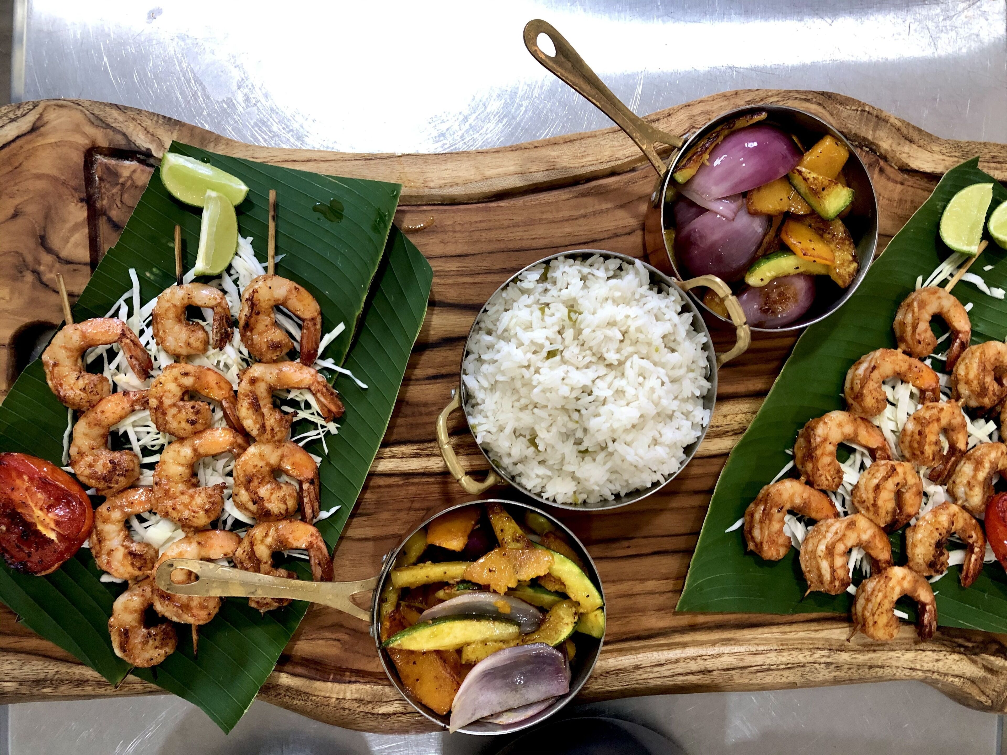 Una bandeja de madera con brochetas de camarones, arroz blanco y un tazón de verduras mezcladas.