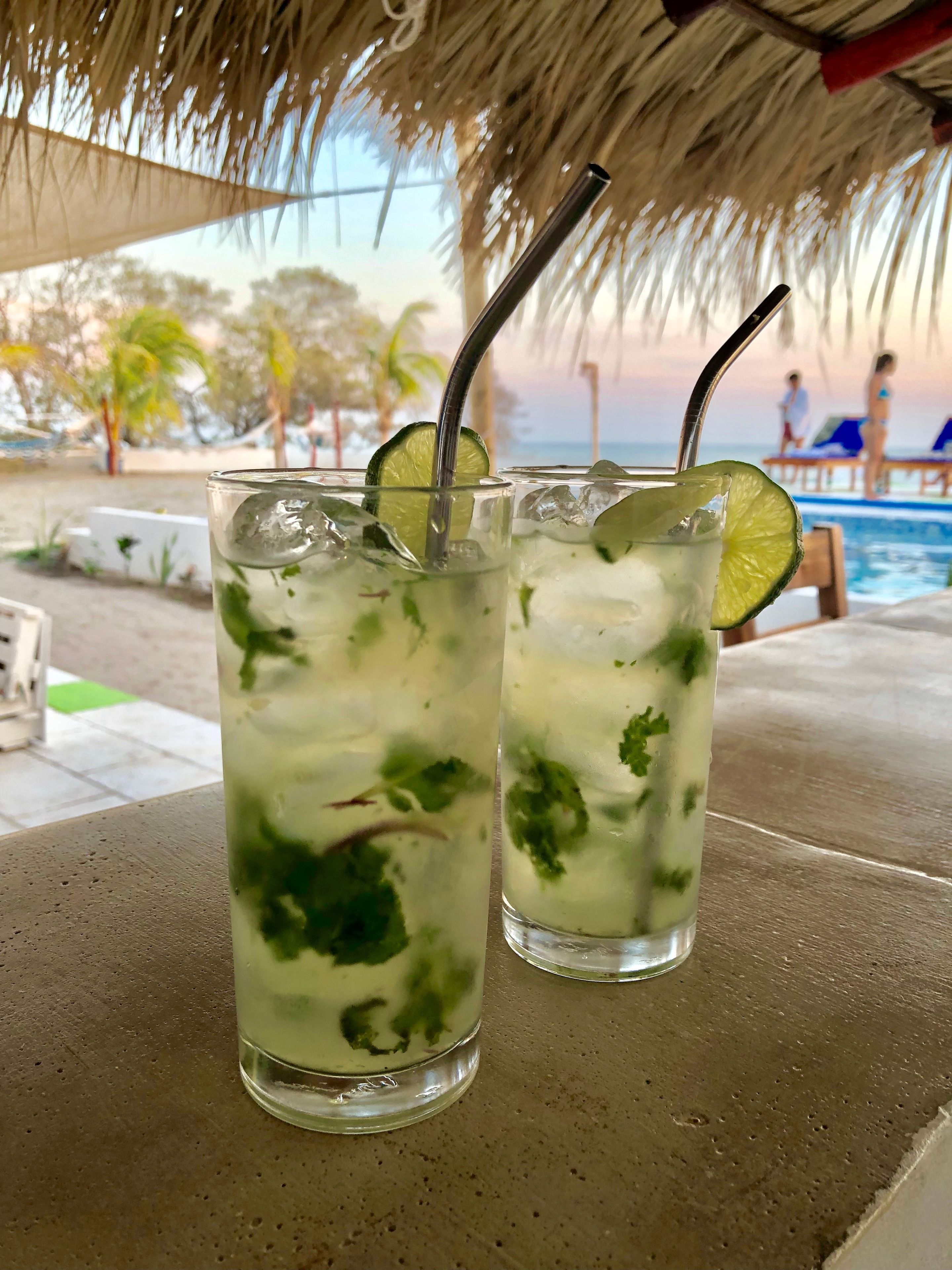 Two glasses of a lime-based drink with mint, placed on a table under a thatched roof.