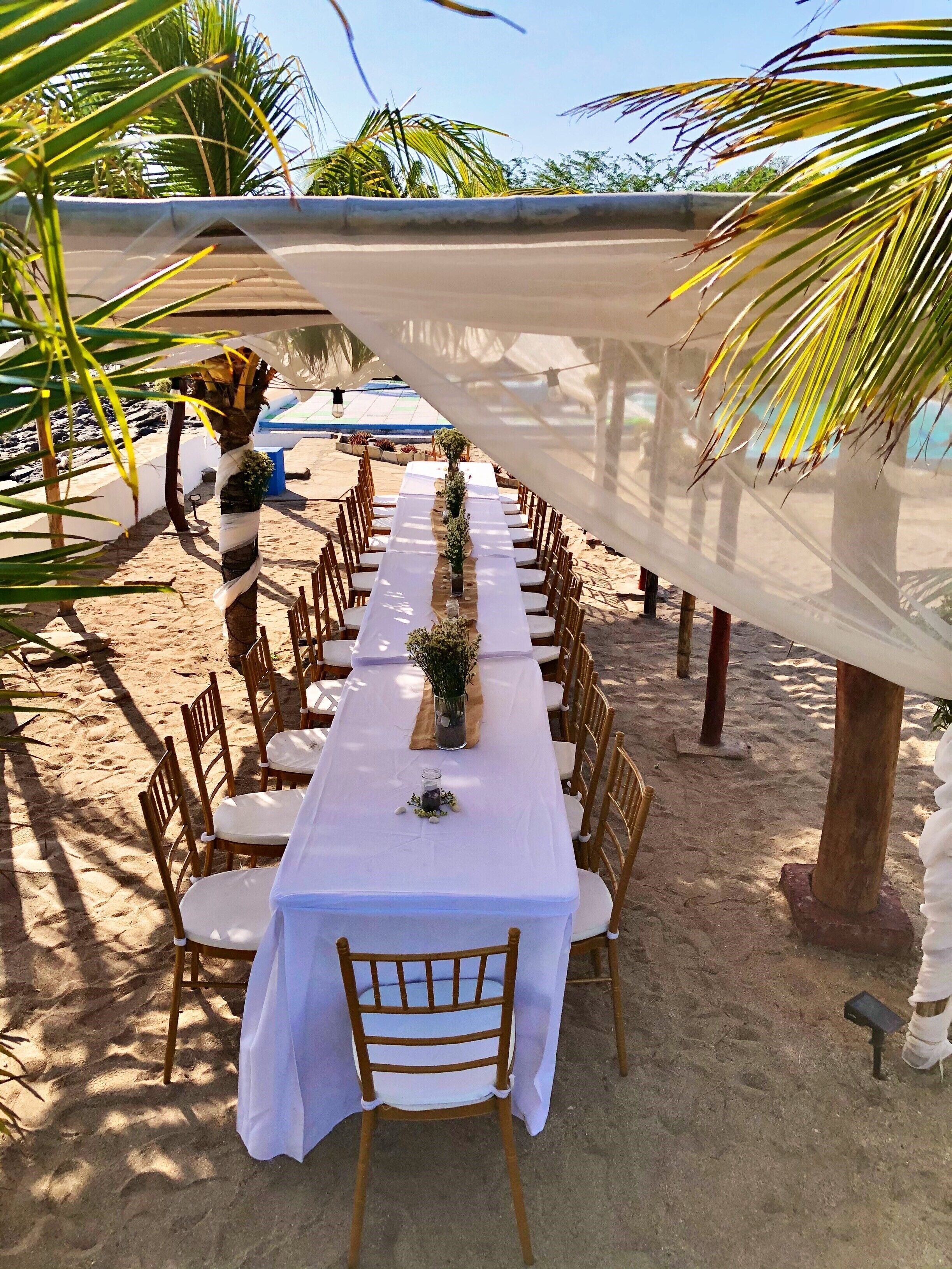 Un montaje para comer frente al mar con una larga mesa blanca, sillas de madera y un toldo en la parte superior.