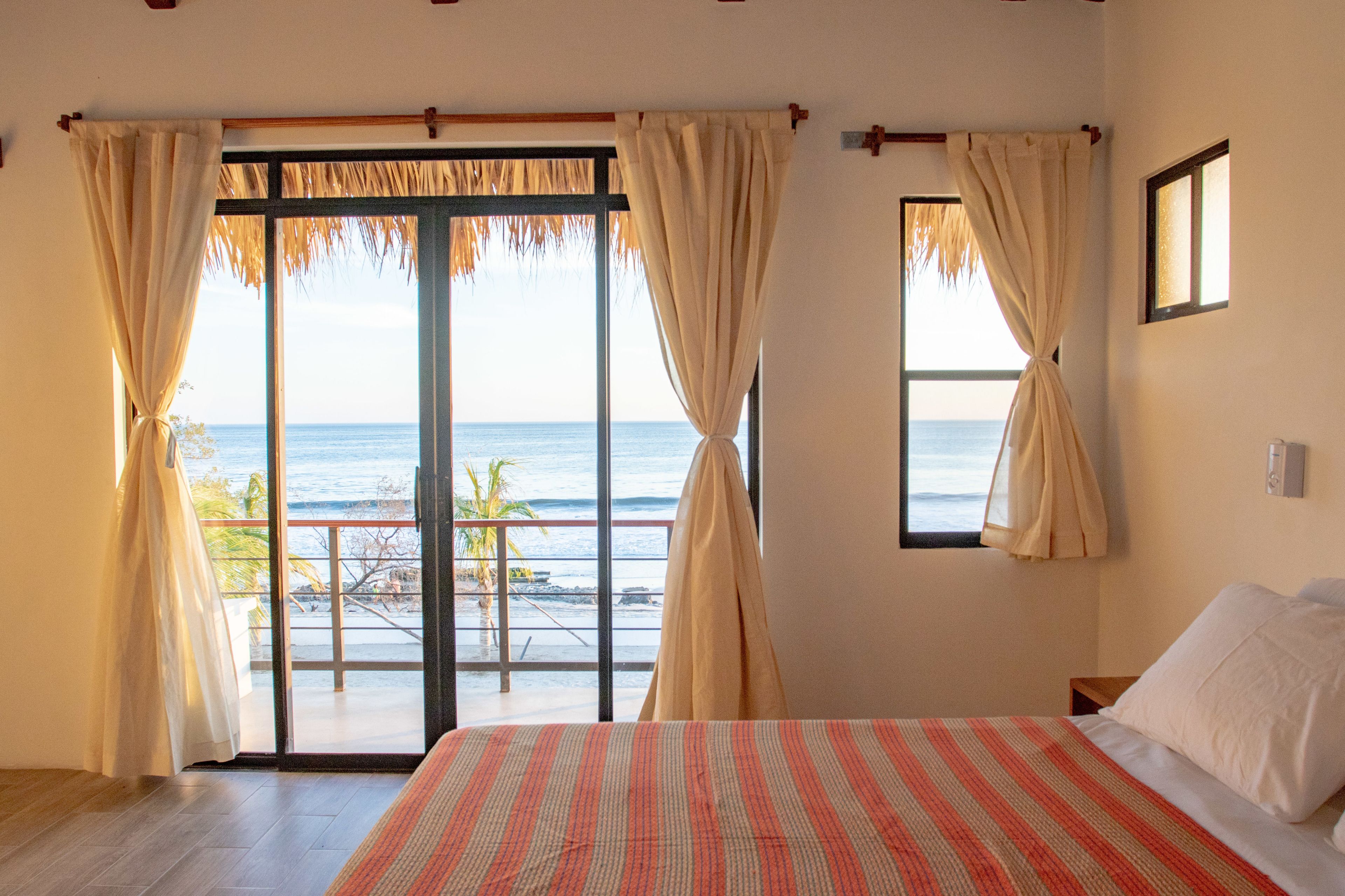 A bedroom with a large bed, a view of the ocean through a sliding glass door, and a balcony with a thatched roof.