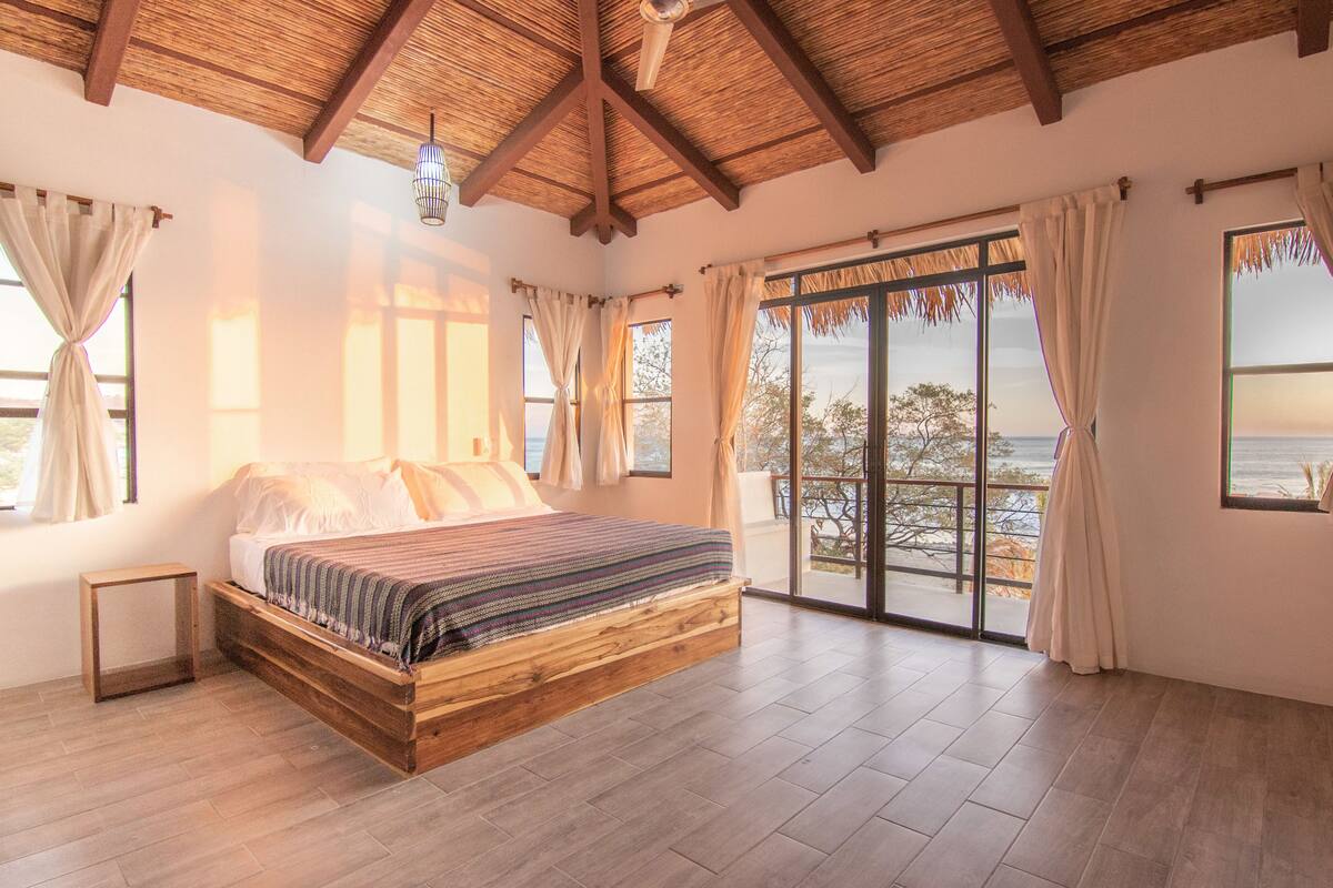 A spacious bedroom with a large bed, wooden beams, and a view of the ocean through large windows.