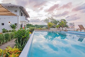 A swimming pool with a thatched roof building, surrounded by tropical plants and a clear sky. - Costa Oeste (Popoyo)