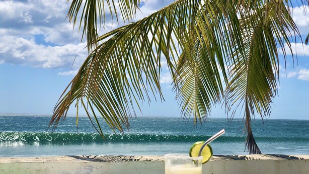 A glass of drink with a lime garnish on a wooden table, palm tree, ocean waves, and a clear sky. - Costa Oeste (Popoyo)