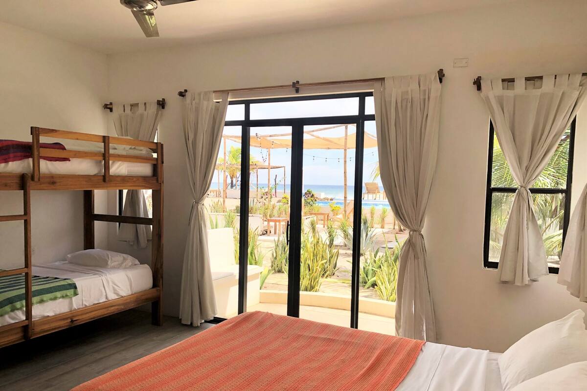 A room with a bunk bed, a single bed with an orange blanket, and a view of a beach and pool through large windows.
