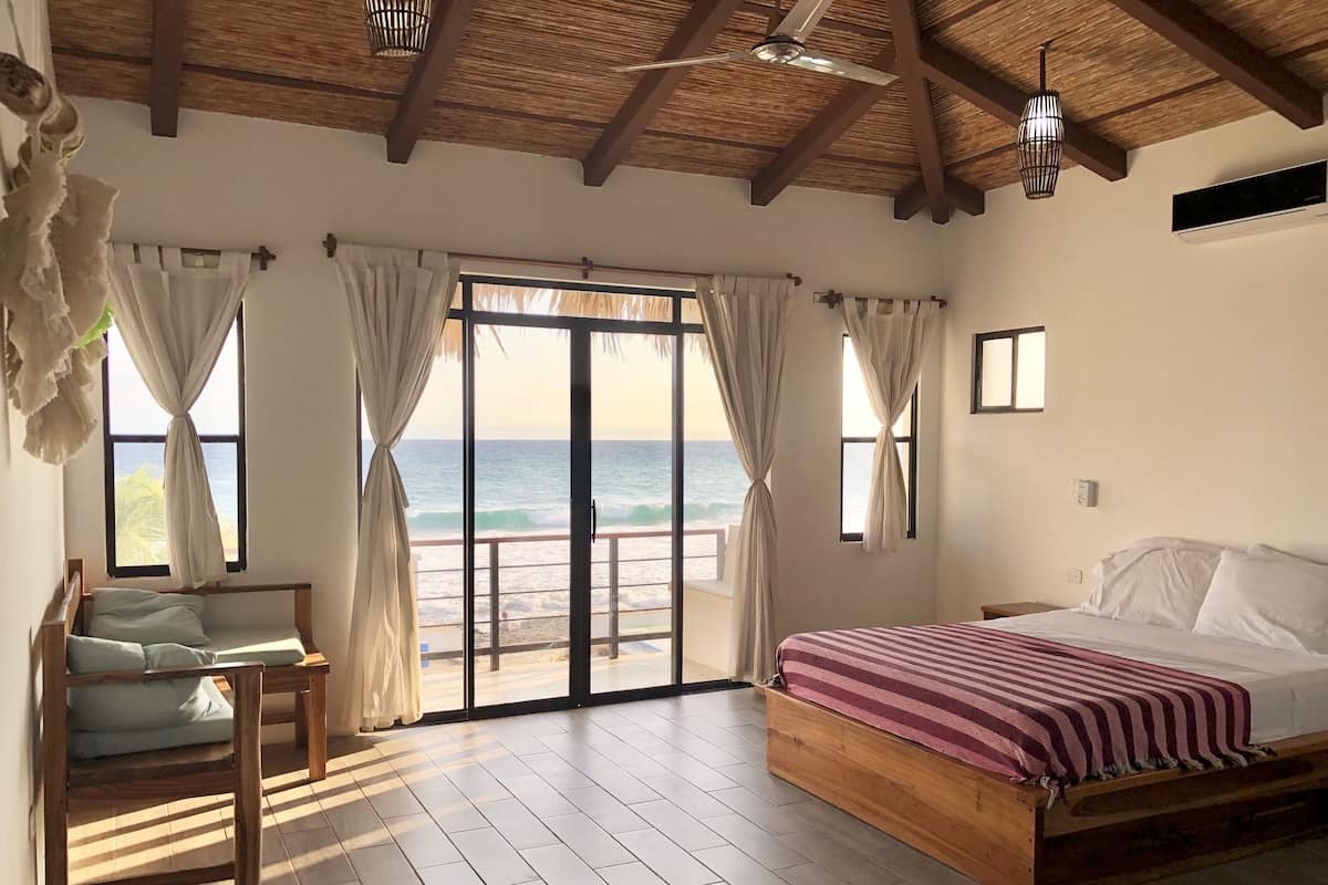 A bedroom with a wooden bed, a view of the ocean through large sliding doors, and a ceiling fan.