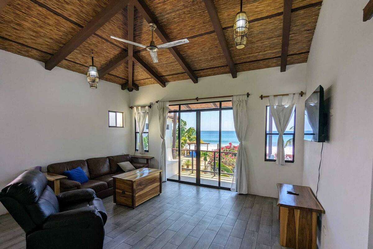 A spacious living room with a wooden ceiling, a leather sofa, a wooden coffee table, and a view of the beach through large sliding glass doors.