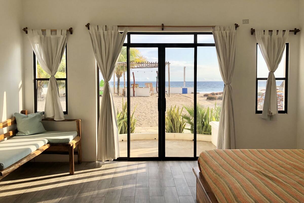 A room with a bed, a sofa, and a large sliding glass door leading to a beach view.
