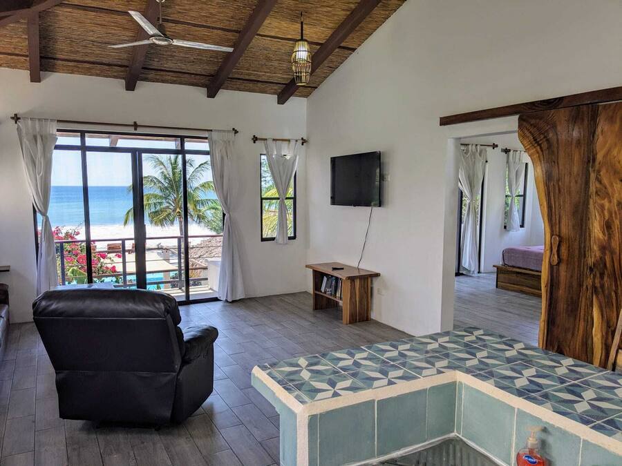 Ein Zimmer mit großem Fenster zum Strand, Deckenventilator, Wand-Fernseher und ein schwarzer Ledersessel.
