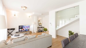 Una sala moderna con un sofá, una mesa de centro y un televisor. Se ve una escalera a la derecha y una planta en el suelo.