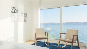 Una habitación con un ventanal grande que da a una masa de agua, dos sillas, una cama y una alfombra azul.
