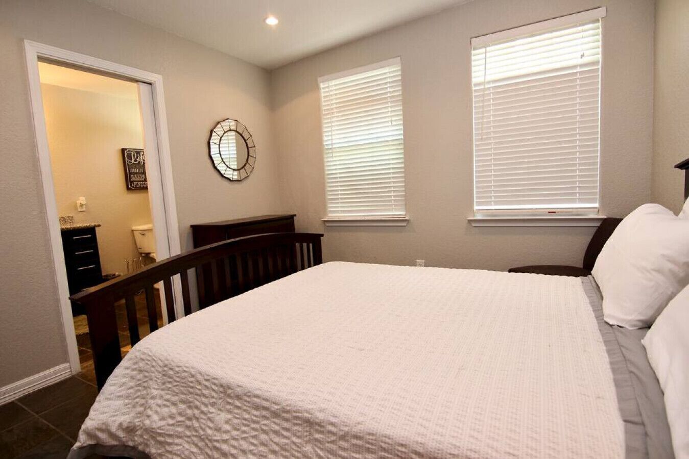 A bedroom with a large bed, two windows with blinds, a wooden headboard, and a small wall decoration.