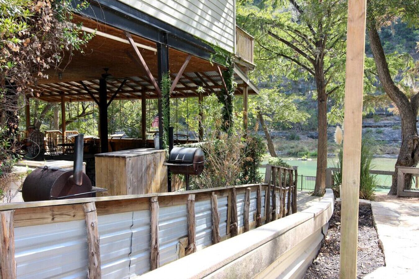 An outdoor patio with a grill and seating area, surrounded by trees and a view of a body of water.