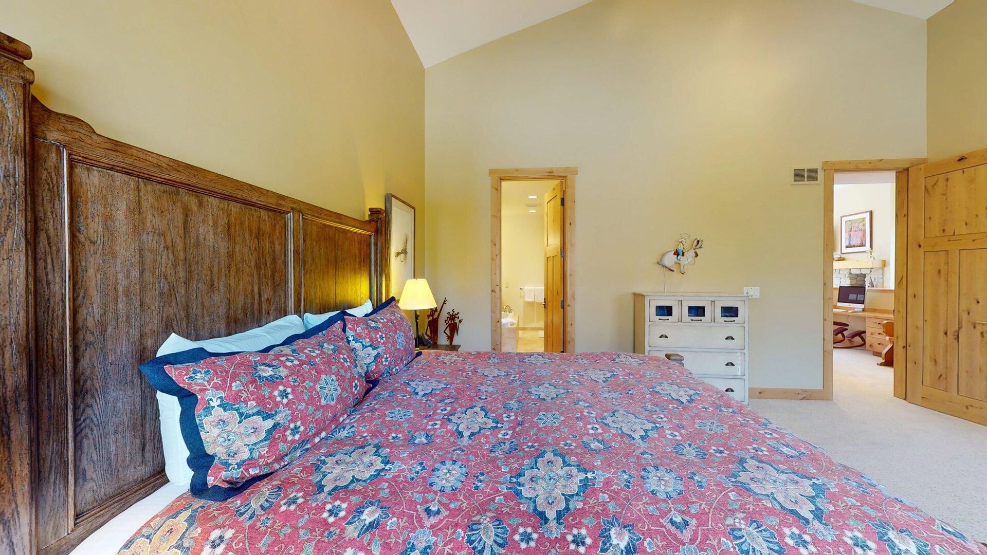 Une chambre à coucher avec un grand lit, une tête de lit en bois, une table de chevet avec une lampe, une commode et une porte donnant sur une autre pièce.