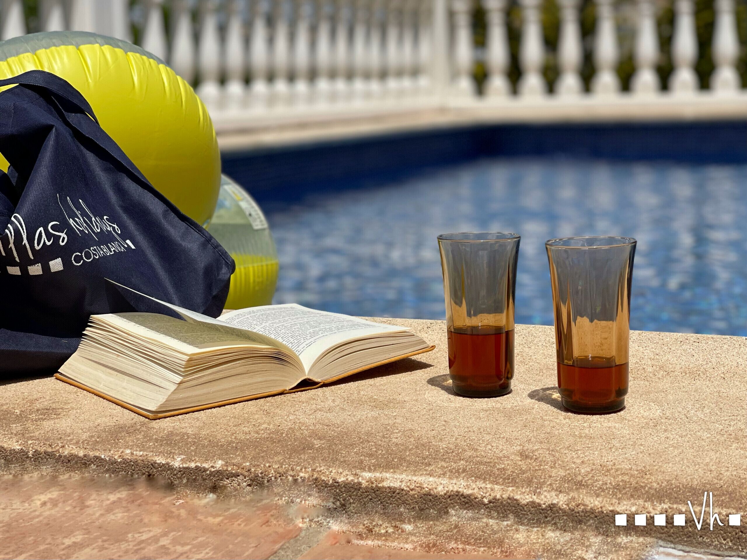 A poolside setting with a book, two glasses of tea, and a yellow beach ball.