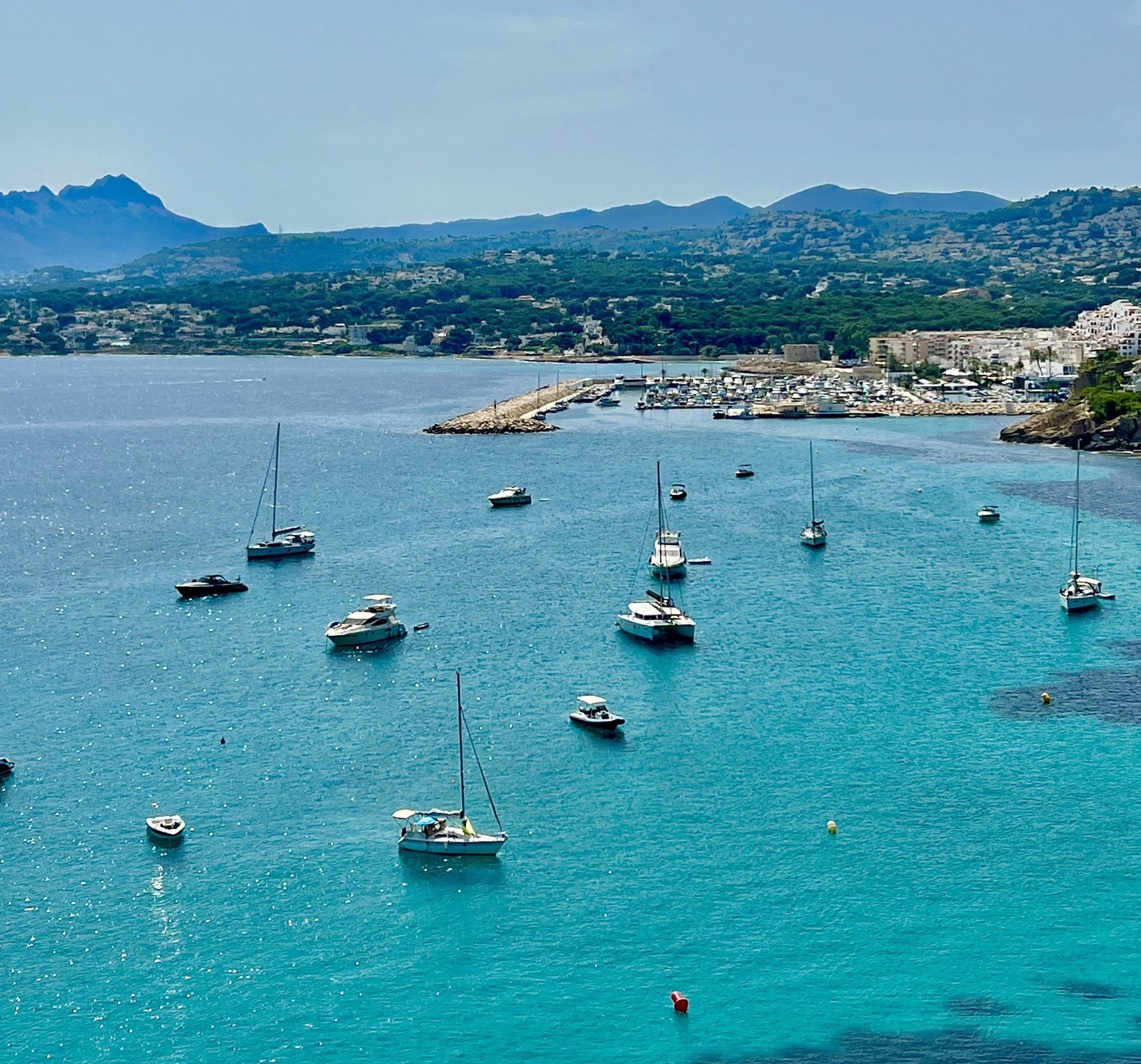 Un puerto deportivo costero con varias embarcaciones fondeadas en aguas tranquilas, rodeado de un paisaje montañoso y un pueblo pequeño al fondo.