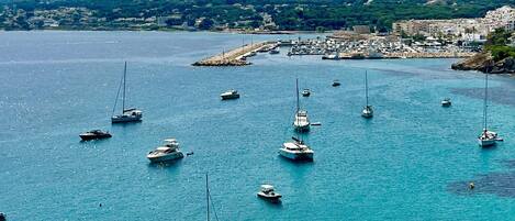 Un puerto deportivo costero con varias embarcaciones fondeadas en aguas tranquilas, rodeado de un paisaje montañoso y un pueblo pequeño al fondo.
