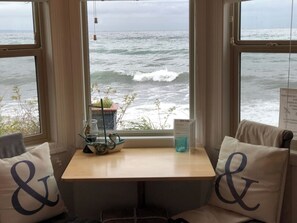 A room with a view of the ocean through large windows, a small table with a hot tub sign, and two chairs with decorative pillows. - Beachfront duplex Cottage (Qualicum Beach)