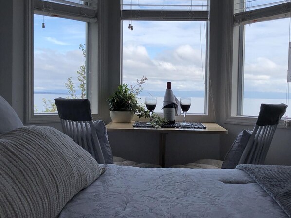 A room with a bed, two chairs, a table with a plant and wine glasses, and a view of the sea through large windows.
