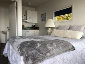 A bedroom with a bed, a bedside lamp, a window with a view, a kitchen area with cabinets, and a sink. - Beachfront duplex Cottage (Qualicum Beach)