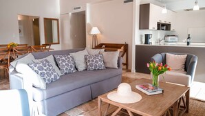 En moderne stue med en grå sofa, mønstrede puder, et træ sofabord med en bog og en hat, og udsigt til et køkkenområde.