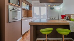 Una cocina con horno empotrado, microondas y ventana con vistas. Hay dos taburetes verdes y una tetera roja sobre la encimera.