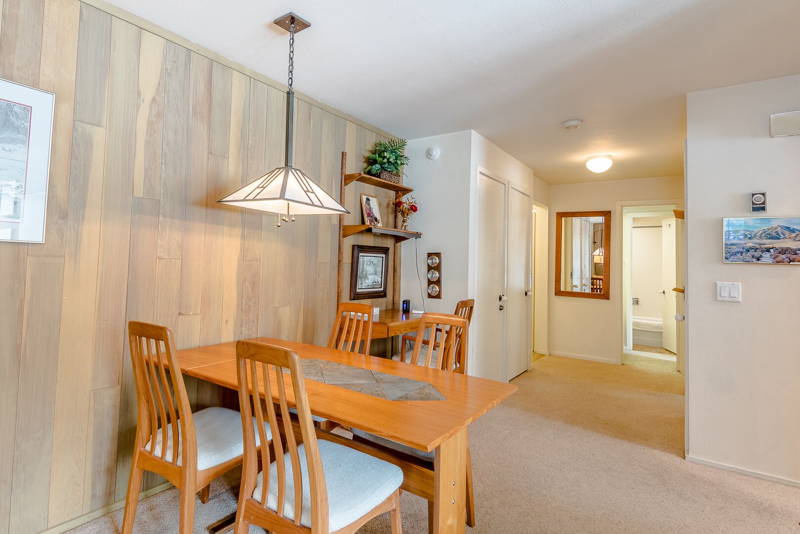 A dining area with a wooden table and chairs, a hanging light fixture, and a hallway leading to other rooms.