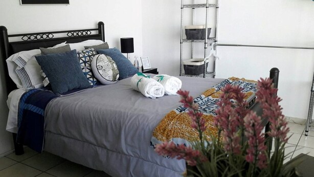 Una cama bien hecha con edredón gris, varios almohadones y una manta estampada. Hay una mesita de noche con una lámpara y un cuadro enmarcado. Una estantería metálica está contra la pared, y una planta en maceta está en primer plano.