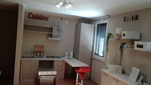 A compact kitchen with a gas stove, a sink, and a countertop. There is a window with blinds, a small plant, and a shelf with storage boxes.
